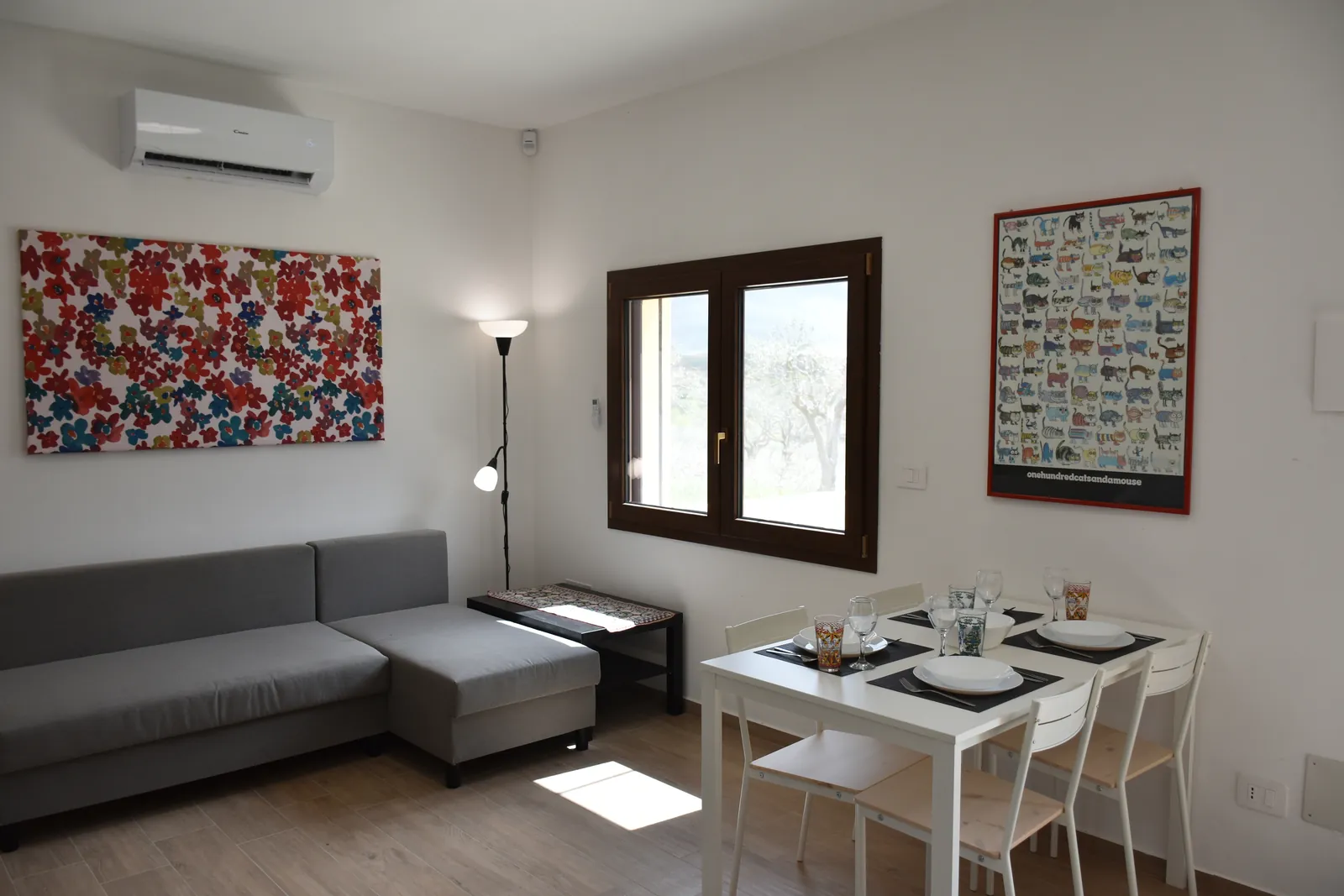 Il soggiorno: divano grigio, quadro a fiori e tavolo bianco apparecchiato per quattro